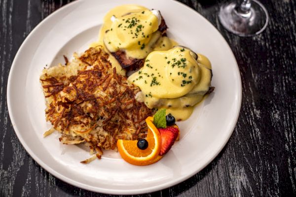 The image shows a plate of Eggs Benedict with hollandaise sauce, hash browns, and garnished fruits including orange slice, strawberry, and blueberries.